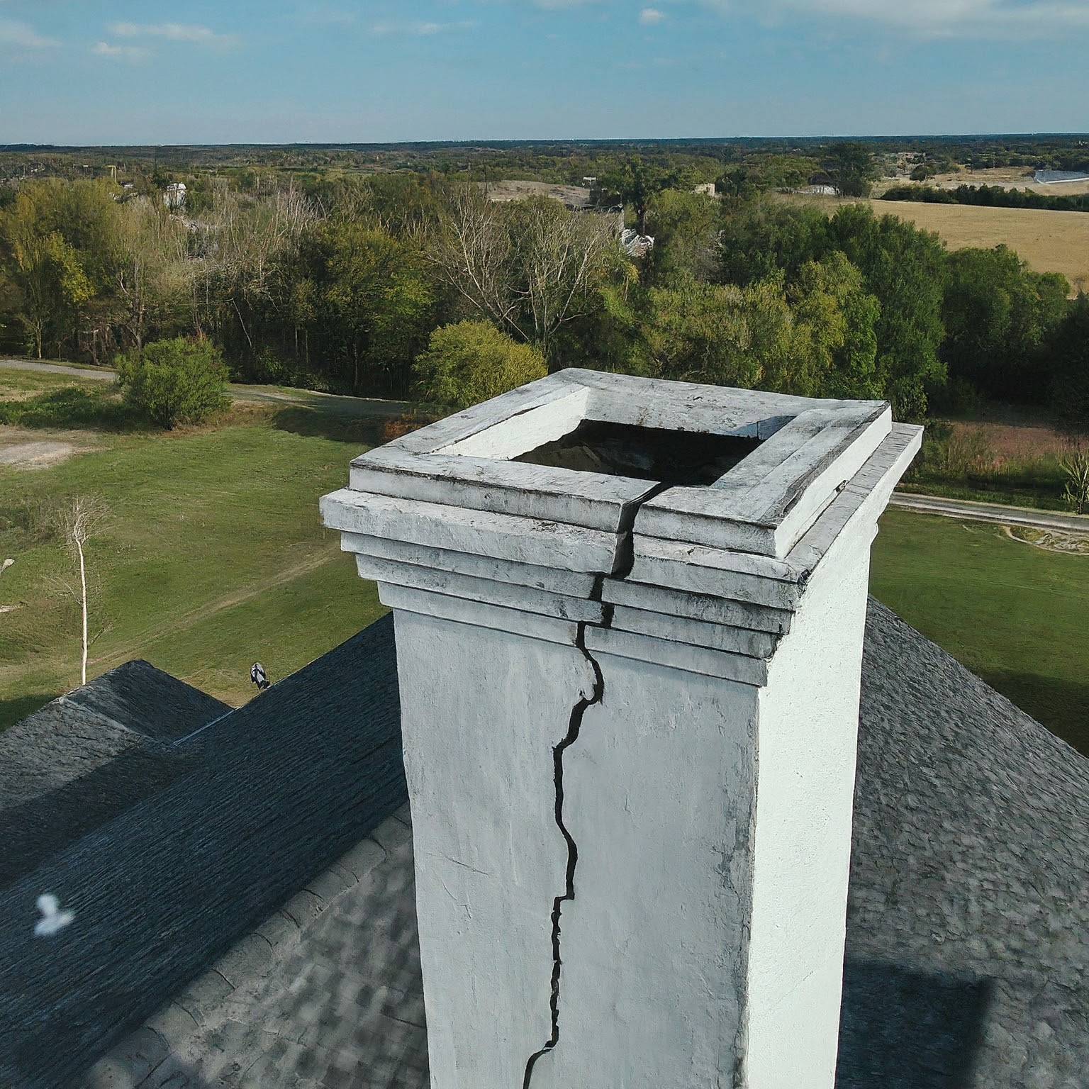white chimney on a rooftop. Several hairline cracks spiderweb across the surface, barely visible from ground level. || Alt attribute: Aerial view of a large white chimney on a rooftop. Hairline cracks are visible on the chimney surface