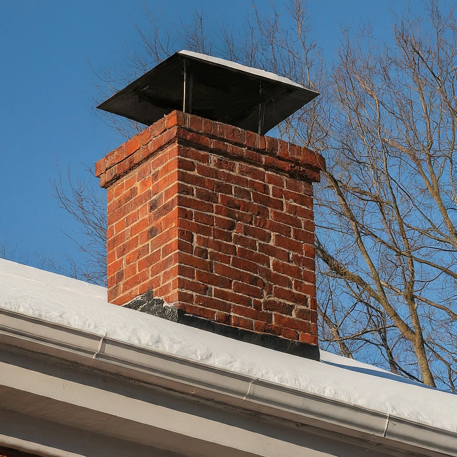 A brick chimney with a metal cap sits on a pitched roof covered in snow - Contact us.