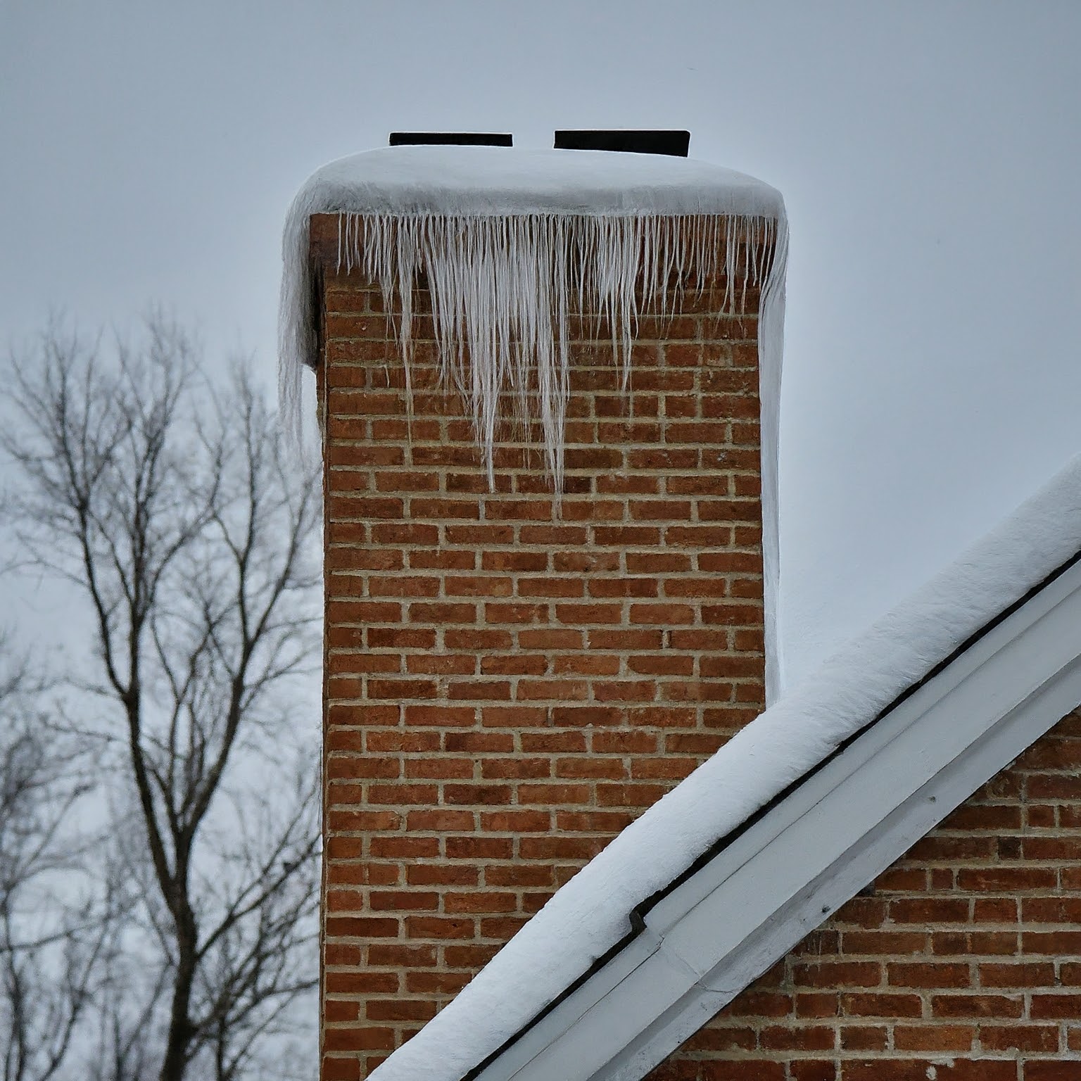  The icicles are long and pointed, and they are hanging from the bottom of the chimney. The bricks of the chimney are red and appear to be old. There is also a metal band around the chimney, near the top.